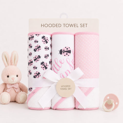 A pink and white hooded towel set in a clear plastic package with a cardboard header, tied with a white ribbon and a round tag. The set includes three towels: one with black ladybugs on a white background, one with pink script text, and one with pink polk