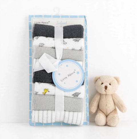 A packaged set of folded baby socks in dark gray, white, and light gray, tied with a white ribbon and a round tag, displayed next to a small beige teddy bear on a white background. The packaging is light blue with a geometric pattern and reads 'Little Apa