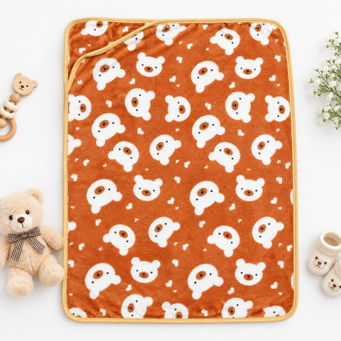 An orange baby blanket with a white bear face pattern, styled in a flat-lay with a teddy bear, teething ring, and baby booties on a white background.