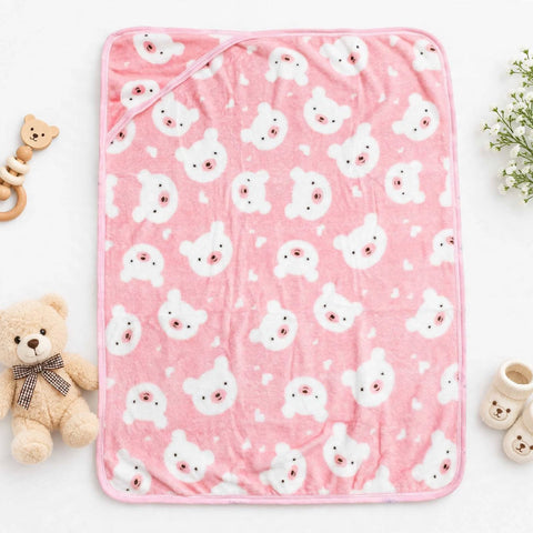 A pink baby blanket with a repeating white bear face pattern, styled in a flat-lay with a teddy bear, baby booties, and a wooden teething ring on a white background.