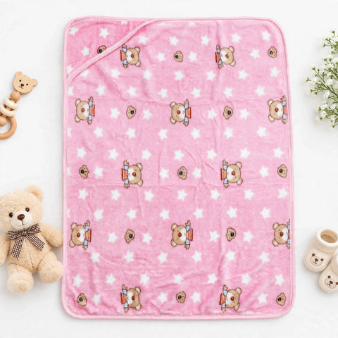 A pink plush blanket with a repeating pattern of teddy bears wearing orange bibs and white stars, styled in a flat-lay with a teddy bear, teething ring, and baby booties on a white background.