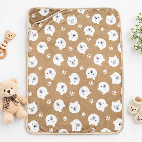 A rectangular baby blanket or mat with a tan background and an allover print of white cartoon bear faces and paw prints, styled in a flat lay with a teddy bear, rattle, and baby booties nearby.
