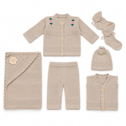 A coordinated baby knit clothing set laid out on a white background, including a cardigan with small floral embroidery, pants, a folded blanket with a flower applique, a hat, a sleeveless vest, and a pair of booties. The items are all in a warm light beig