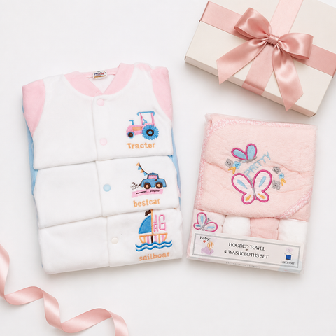 A baby gift set featuring a white and pink baby cardigan with embroidered tractor, car, and sailboat designs, next to a pink plush hooded towel and matching packaged towels. A cream gift box with a pink satin bow sits in the background on a white surface.