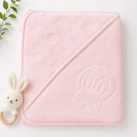 A folded light pink hooded baby towel with a bunny face embossed on one corner, shown on a white background with a small plush bunny teether and green leaves as props.