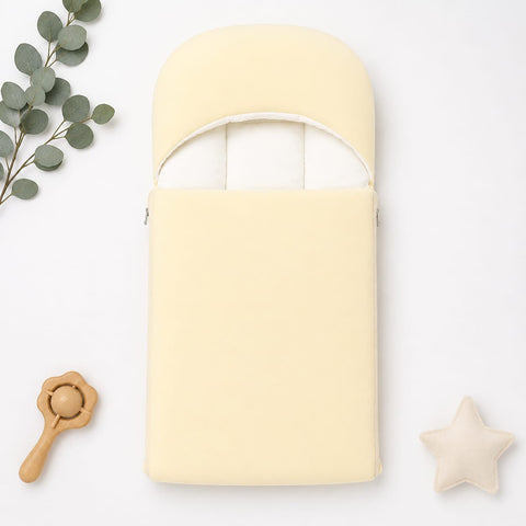 A pale yellow baby footmuff or swaddle pouch with a curved opening, shown in a styled flat-lay on a white background with eucalyptus leaves, a wooden rattle, and a cream star toy.