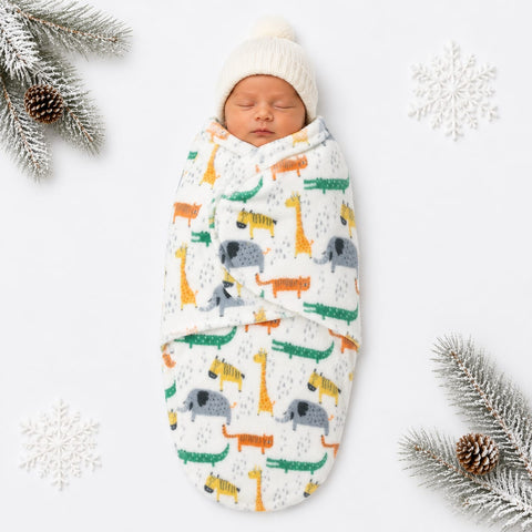 A sleeping newborn baby swaddled in a white fleece blanket with a colorful animal print of giraffes, elephants, crocodiles, and zebras, paired with a cream knit beanie hat. The scene is styled with white snowflakes and pine branches on a white background.