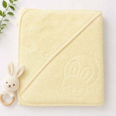 A pale yellow hooded baby blanket with a bunny face motif, styled flat-lay with a small plush bunny teething ring toy on a white background.