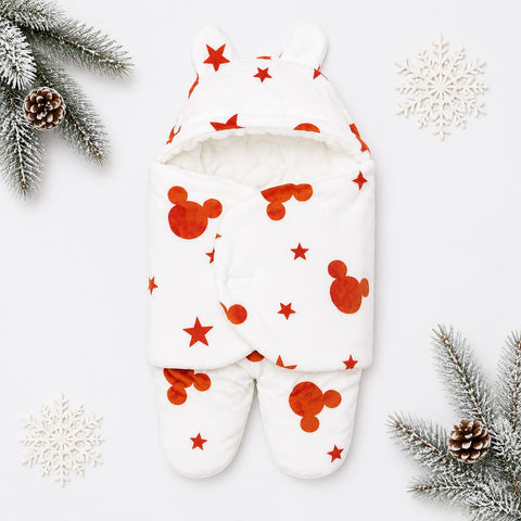 A baby footed swaddle blanket with a hood and ear details, featuring a white background with red Mickey Mouse head silhouettes and red stars. The swaddle is styled in a flat-lay with winter-themed props including pine branches, pinecones, and white snowfl