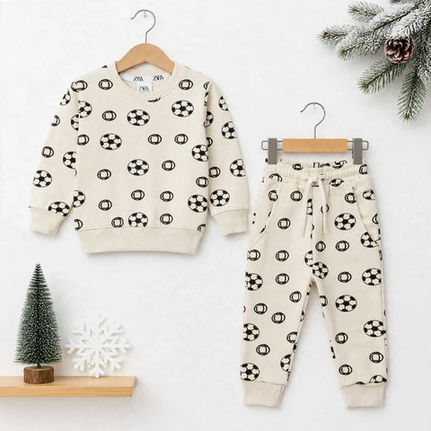 A children's two-piece outfit set consisting of a long-sleeve sweatshirt and matching jogger pants, both in a warm cream color with a black soccer ball pattern, displayed on wooden hangers against a white background with holiday props.