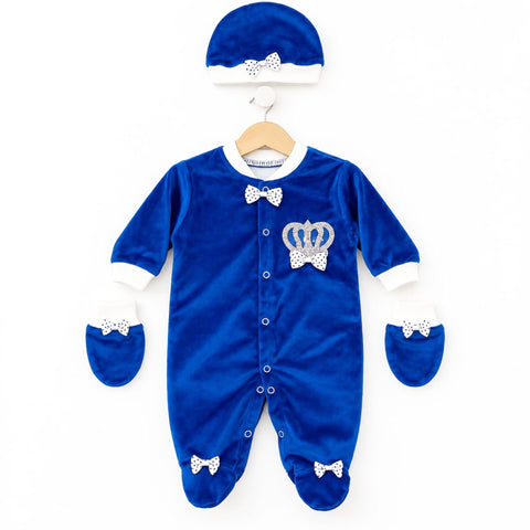 A royal blue velvet baby outfit set including a footed one-piece sleeper, matching booties, and a bonnet hat, all decorated with white polka-dot bow details and a silver crown applique on the chest, displayed on a wooden hanger against a white background.
