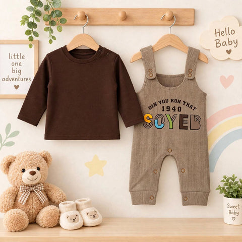 A baby clothing set displayed on wooden hangers against a decorated wall, featuring a dark brown long-sleeve top and a taupe-beige overall jumpsuit with colorful 'SOYEB' lettering. A teddy bear, small booties, and nursery wall decor are arranged around th