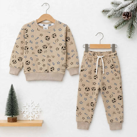 A children's two-piece outfit set consisting of a beige long-sleeve sweatshirt and matching jogger pants, both decorated with an all-over print of black soccer balls, blue stars, and brown scribbles. The sweatshirt is shown on a wooden hanger on the left,