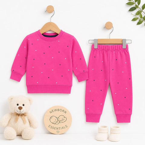 A baby clothing set displayed on wooden hangers against a white background, featuring a bright pink long-sleeve sweatshirt and matching jogger pants with small scattered polka dots. A cream teddy bear sits at lower left, a round wooden sign reading 'Newbo