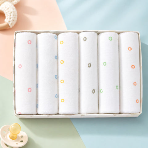 Six rolled white baby blankets or swaddle cloths with small multicolor flower prints, displayed in a rectangular gift box on a pastel-colored surface with baby props.