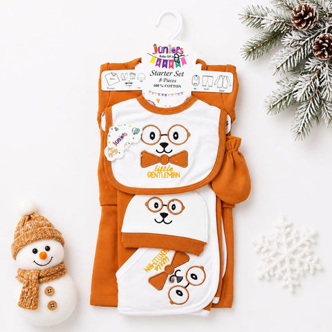 A baby starter set featuring an orange and white knit outfit with matching bibs and a small mitt, displayed on a hanger against a white background with holiday props including a snowman, pine branches, and a snowflake.