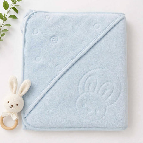 A light blue hooded baby towel with an embossed bunny face on one corner, laid flat on a white background next to a small plush bunny teething ring toy with a wooden ring.