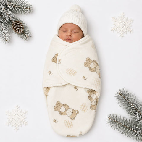 A sleeping newborn baby wrapped in a cream-colored swaddle blanket with a teddy bear pattern, wearing a matching cream knit hat, styled with pine branches and white snowflake decorations on a white background.