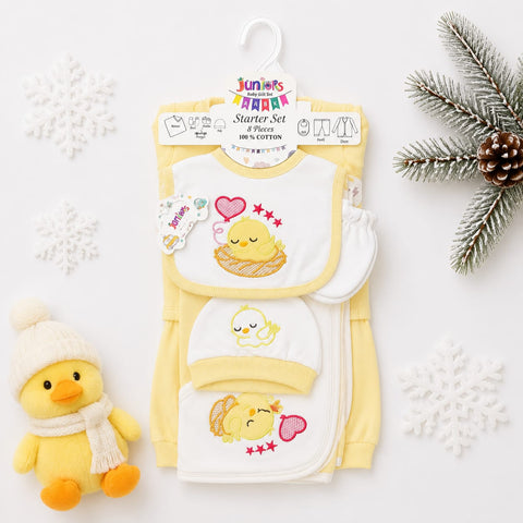 A baby starter set in soft yellow and white, featuring a long-sleeve romper, bib, and footie mitt with cute yellow duck and chick illustrations, displayed on a hanger with decorative snowflakes, a plush duck, and a pine branch.
