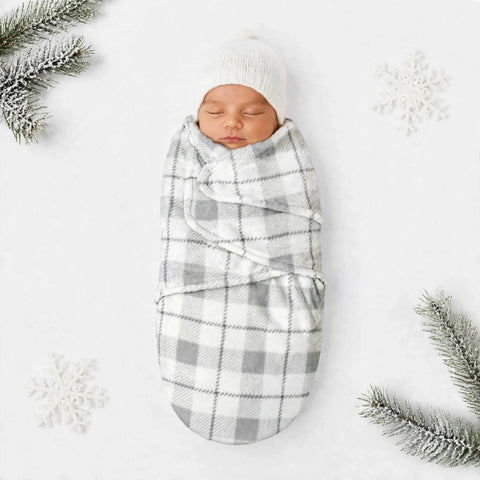 A sleeping newborn baby swaddled in a soft gray and white plaid blanket, wrapped in a white knit hat, laid on a white background with decorative snowflakes and pine branches.