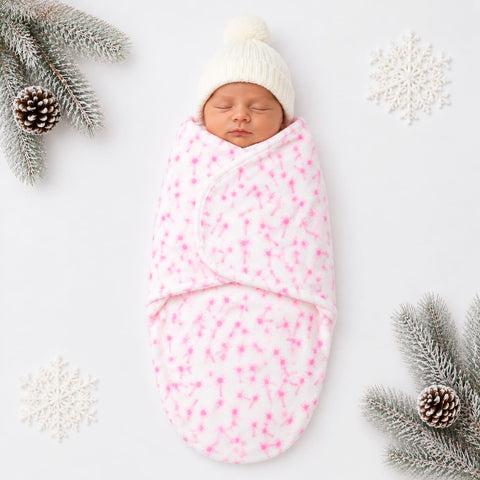 A sleeping newborn baby wrapped in a white plush swaddle blanket with a pink star pattern, wearing a white knit beanie, styled with winter-themed props on a white background.