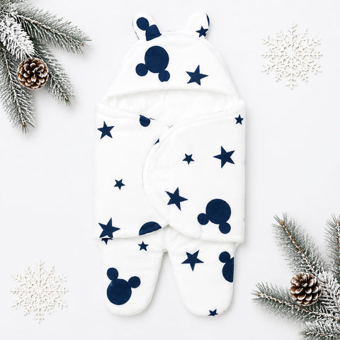 A white footed baby swaddle blanket with a navy blue Mickey Mouse silhouette and star pattern, styled in a flat lay with pine branches, snowflakes, and pinecones on a white background.