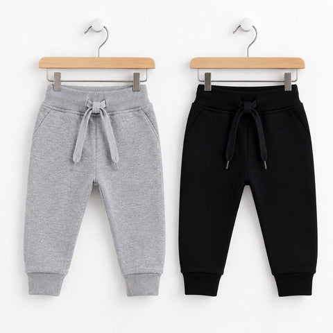Two pairs of children's jogger sweatpants hanging on wooden hangers against a white background. The pair on the left is light heather gray with a matching drawstring, and the pair on the right is solid black with a matching drawstring.