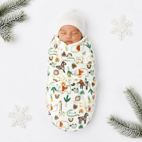 A sleeping newborn baby wrapped in a cream-colored swaddle blanket with a playful safari animal print, wearing a matching cream knit beanie. The scene is styled with white snowflakes and pine branches on a clean white background.