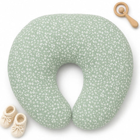 A U-shaped nursing pillow in sage green with a white floral pattern, styled with a wooden baby rattle and cream-colored knit booties on a white background.