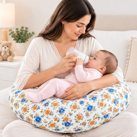 A woman in a cream lace-trim top sits on a white bed holding a baby in a floral nursing pillow while feeding the baby with a bottle. The nursing pillow has a white background with a blue and orange floral pattern.
