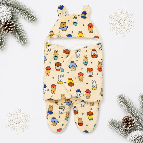 A baby footed swaddle blanket with a hood, featuring a playful multicolor cartoon animal print on a warm cream background. The swaddle is styled in a flat-lay arrangement with winter-themed props including pine branches, pinecones, and white snowflakes on