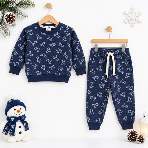 A children's two-piece pajama set hanging on wooden hangers against a white wall, featuring a navy blue sweatshirt and matching joggers with a white bone pattern. The set is styled with winter props including a snowman plush, pinecones, a snowflake orname