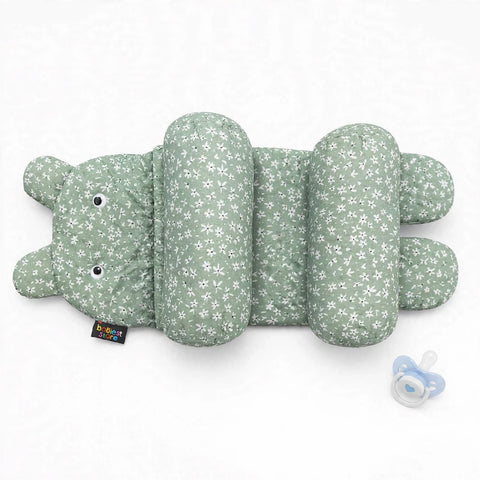 A green and white floral-patterned baby support pillow shaped like a hippo, shown on a white background with a small blue pacifier nearby. A small brand tag is visible on the side.