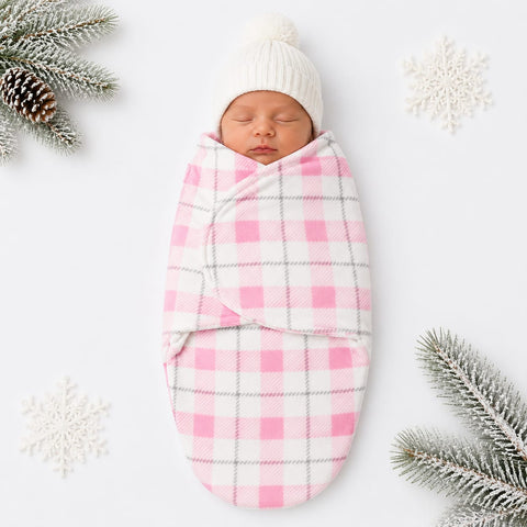 A sleeping newborn baby swaddled in a pink and white gingham blanket with a white knit hat, styled with winter greenery and snowflakes on a white background.