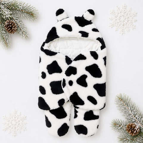 A plush baby footed sleeper or onesie in a black and white cow print pattern, styled flat-lay on a white background with winter-themed decorative props including pine branches, pinecones, and white snowflakes.