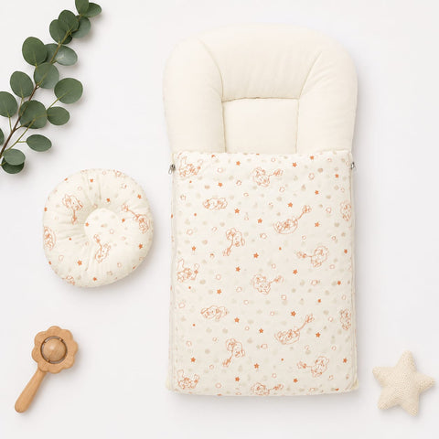 A baby bassinet footmuff and matching circular pillow set in warm cream fabric with a delicate orange bunny and star print, styled on a white background with eucalyptus leaves, a wooden rattle, and a star toy.