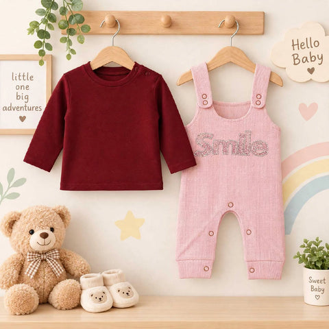 A baby clothing set displayed on wooden hangers against a decorated wall, featuring a long-sleeve crewneck shirt in deep burgundy and matching pink overalls with 'Smile' written across the chest in rhinestones. A teddy bear, small booties, and nursery dec