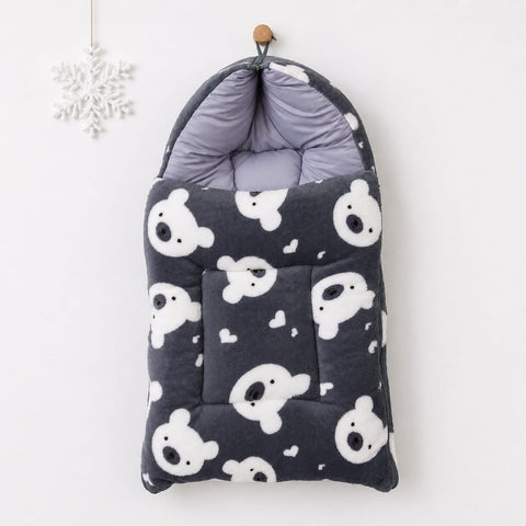 A quilted baby footmuff in dark gray with a white teddy bear pattern, hanging from a wooden peg on a white wall next to a white snowflake ornament.