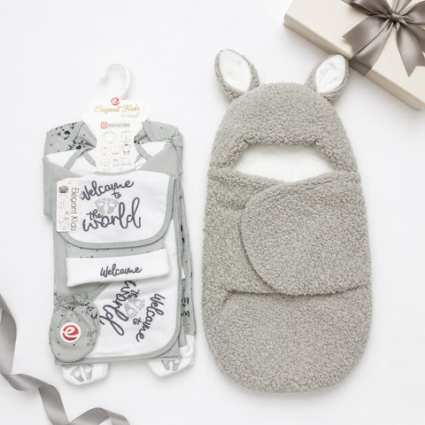 A baby welcome gift set from Organic Kids, featuring a plush gray and white fleece bootie with bunny ears and a matching footie cover, shown next to its retail packaging on a white background with a gift box and ribbon.