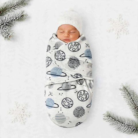 A sleeping newborn baby swaddled in a white wrap blanket with a blue and black space-themed print of planets, moons, and stars, wearing a white knit hat. The baby is photographed from a top-down flat-lay perspective on a white textured background decorate