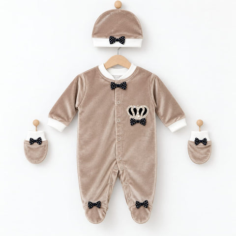 A baby clothing set in taupe velvet, including a footless one-piece jumpsuit, a matching beanie hat, and two booties, all decorated with black and white polka-dot bow ties and a small crown applique on the chest.