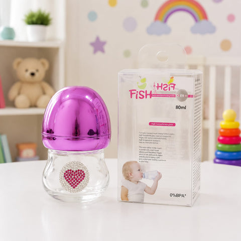 A clear baby bottle with a shiny metallic magenta-purple cap and a rhinestone heart emblem on the front, shown next to its retail box labeled 'Her Fish' on a white tabletop. The background is a softly blurred nursery setting with a teddy bear, shelf, and 