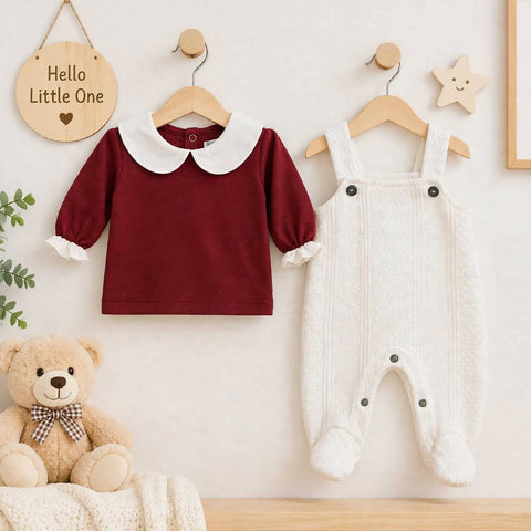 A baby clothing set displayed on wooden hangers against a light wall, featuring a long-sleeve burgundy top with a white Peter Pan collar and white ruffle trim, paired with white textured footless overalls with black buttons. A teddy bear and wooden decor 