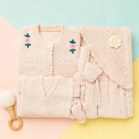 A folded knit baby outfit set in a soft blush pink, including a cardigan with embroidered flowers, a matching wrap or blanket with a flower applique, and a small wooden-handled baby rattle on a pastel geometric background.