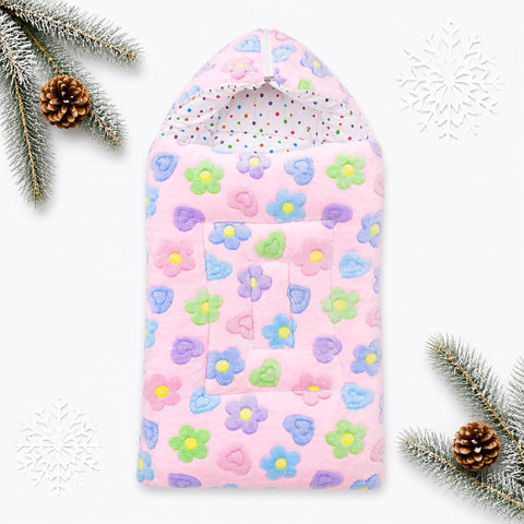 A pink baby blanket or swaddle with a hood, decorated with a colorful pattern of flowers, hearts, and dots, styled on a white background with pine branches, pinecones, and white snowflake decorations.