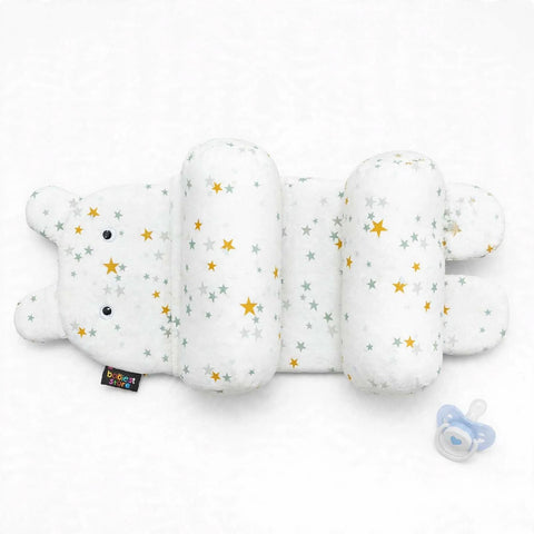 A plush baby support pillow shaped like a caterpillar, featuring a white fabric body with small gray and yellow star patterns. A small black brand tag is visible on the left side, and a blue pacifier lies to the right on a clean white background.