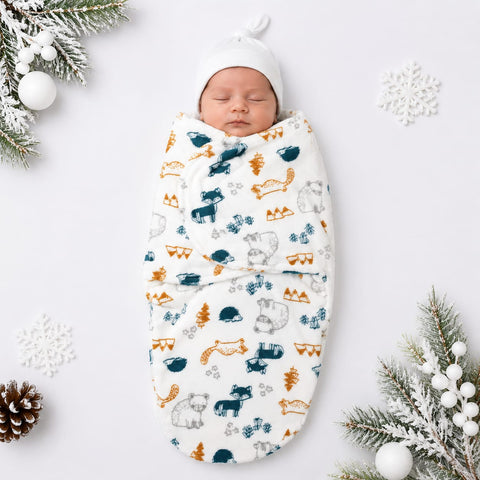 A sleeping newborn baby wrapped in a white swaddle blanket with a colorful forest pattern of moose, foxes, pine trees, and snowflakes, wearing a matching white knit hat. The photo is styled with winter-themed props including pine branches, white ornaments