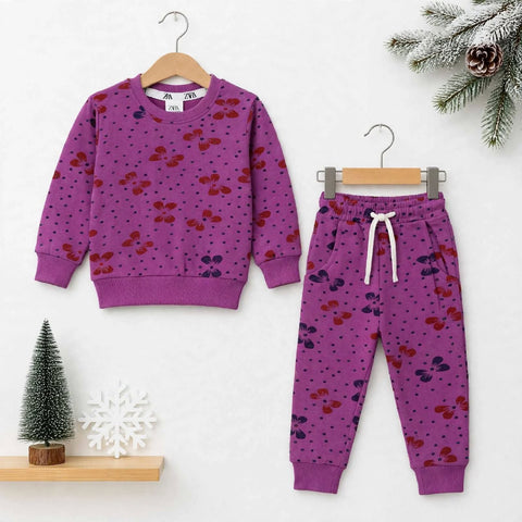 A children's two-piece pajama set featuring a purple long-sleeve top and matching jogger pants with a cherry print, displayed on wooden hangers against a white background with Christmas decorations.