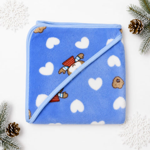A folded blue plush blanket with a diagonal seam, decorated with white hearts, small brown bear faces, and a cute bear peeking out wearing a red striped hat. The blanket is styled in a flat-lay with pine branches and white snowflake decorations on a white