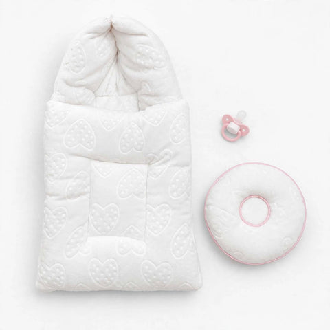 A white quilted baby sleep bag with a hood and embossed heart pattern, shown on a white background with a pink pacifier and a matching pink-trimmed circular baby cushion.